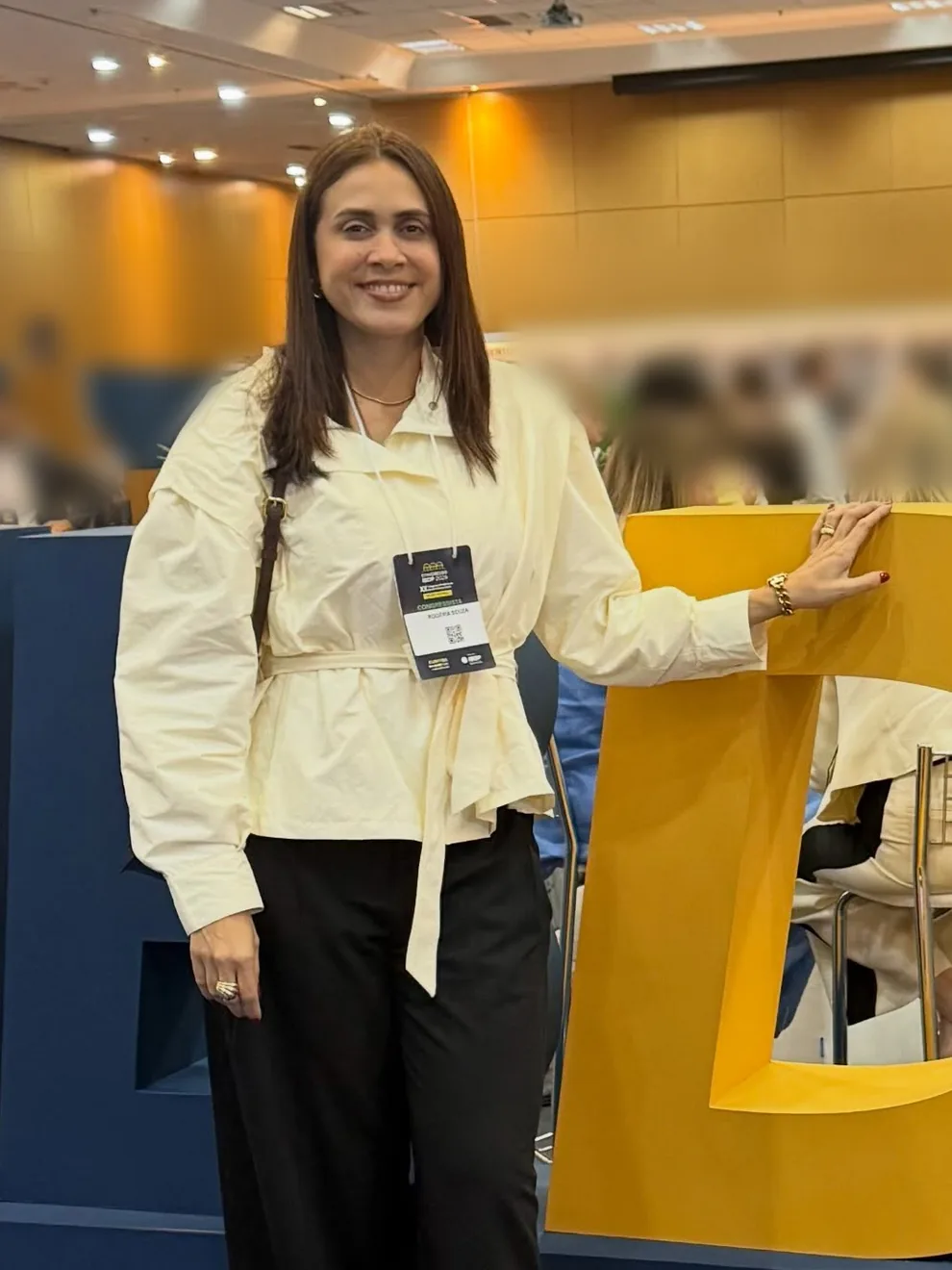 Rogéria Valentim sorrindo, de camisa creme e crachá no pescoço, com a mão apoiada no letreiro amarelo do congresso do IBDP